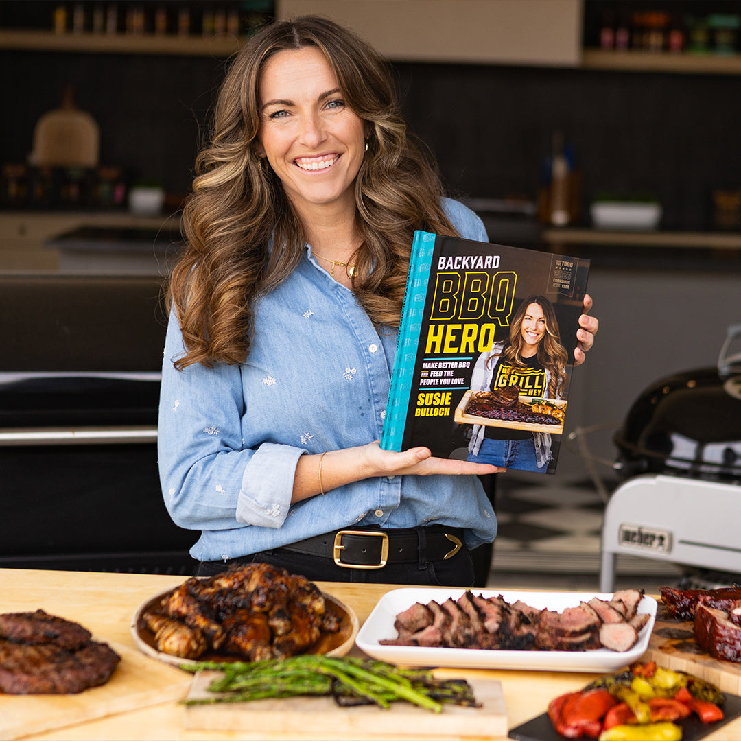 Woman holding a book titled 'Backyard BBQ Hero' in a kitchen with grilled food on a table.