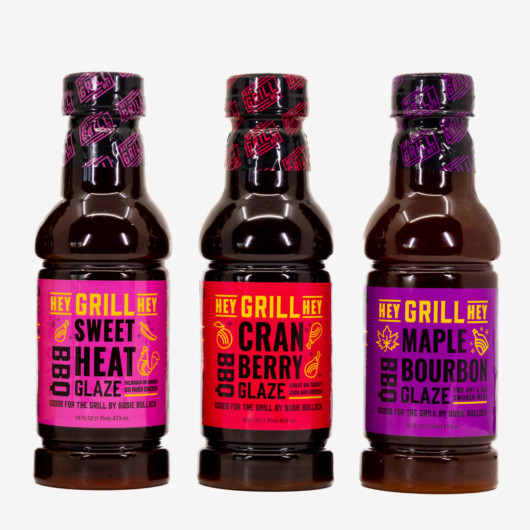 Three bottles of BBQ sauce glaze lined up in a row. The first bottle on the left is Hey Grill Hey Sweet Heat BBQ Glaze, the second bottle says Hey Grill Hey Cranberry BBQ Glaze, and the last bottle is Hey Grill Hey Maple Bourbon Glaze.