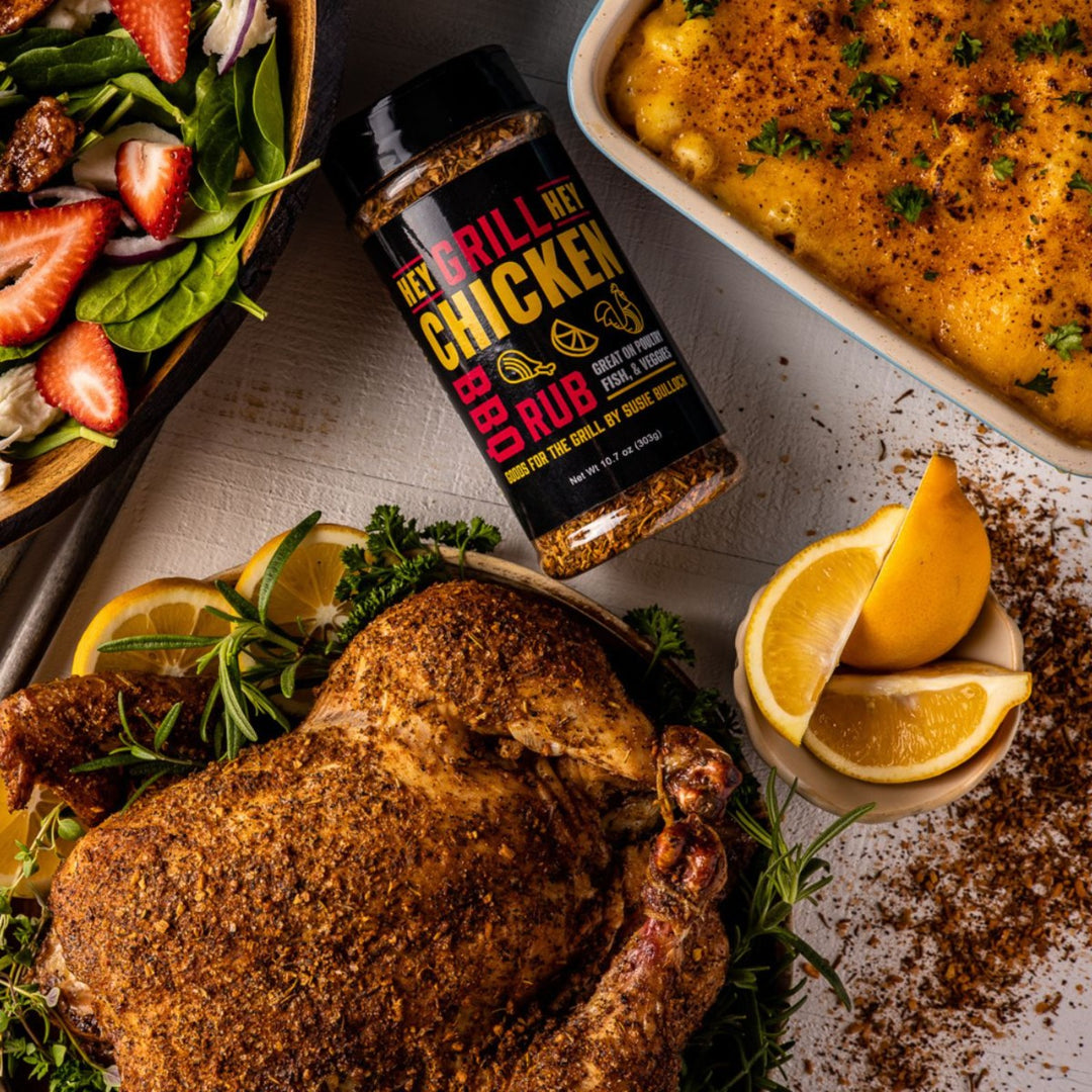 A bottle of Chicken Rub sits on a table with a roasted chicken on a plate with herbs and lemon, and other side dishes. 