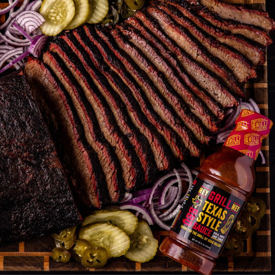 A bottle of texas style sauce sits on a cutting board with sliced smoked brisket and pickles, onions, and jalapenos as garnishing. 