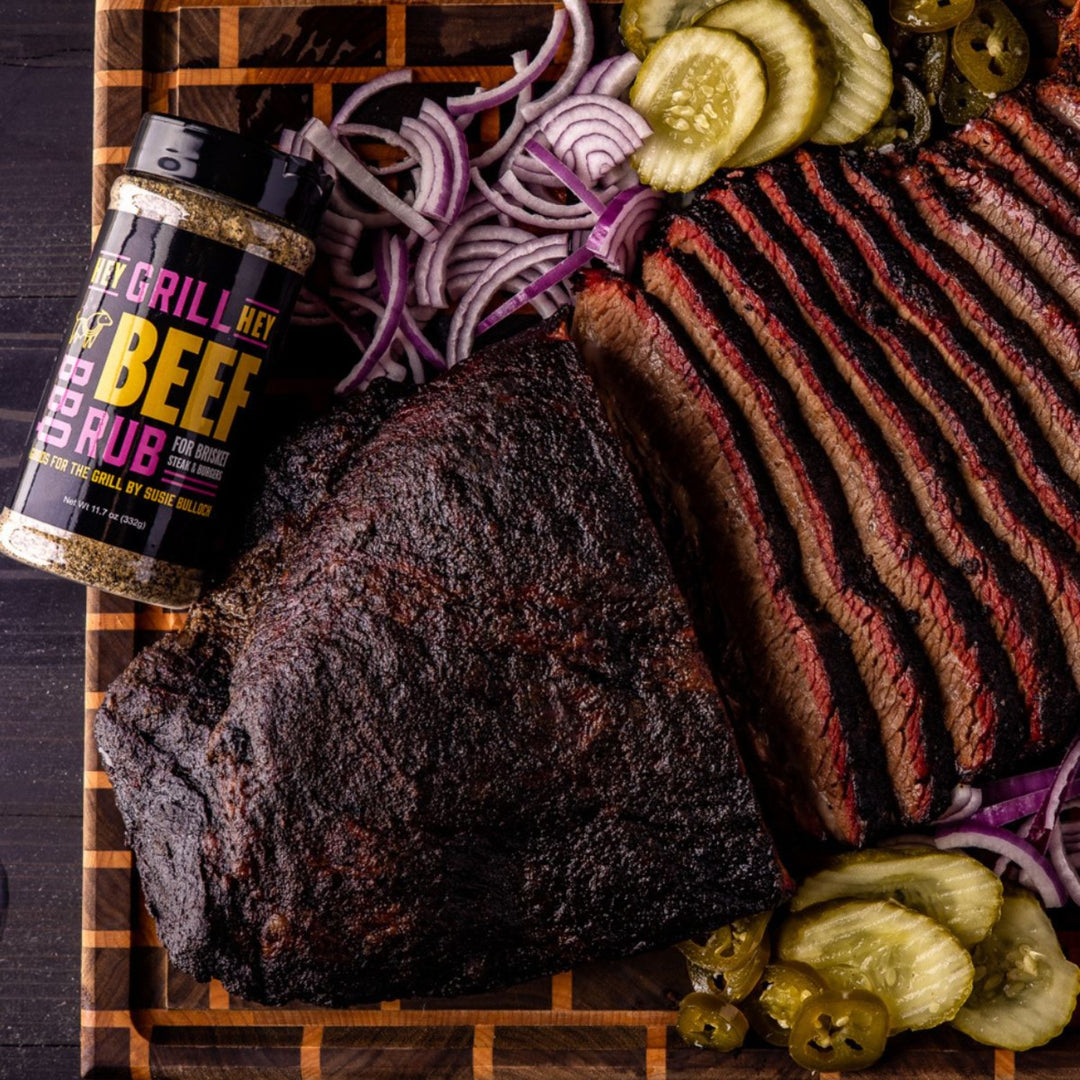 A bottle of Beef Rub sits on a cutting board with sliced smoked brisket and pickles, jalapenos, and onions as garnishing. 