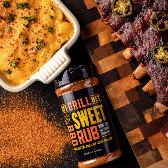 A bottle of sweet rub sits on a table next to a cutting board with BBQ Ribs, and a dish of mac and cheese. 