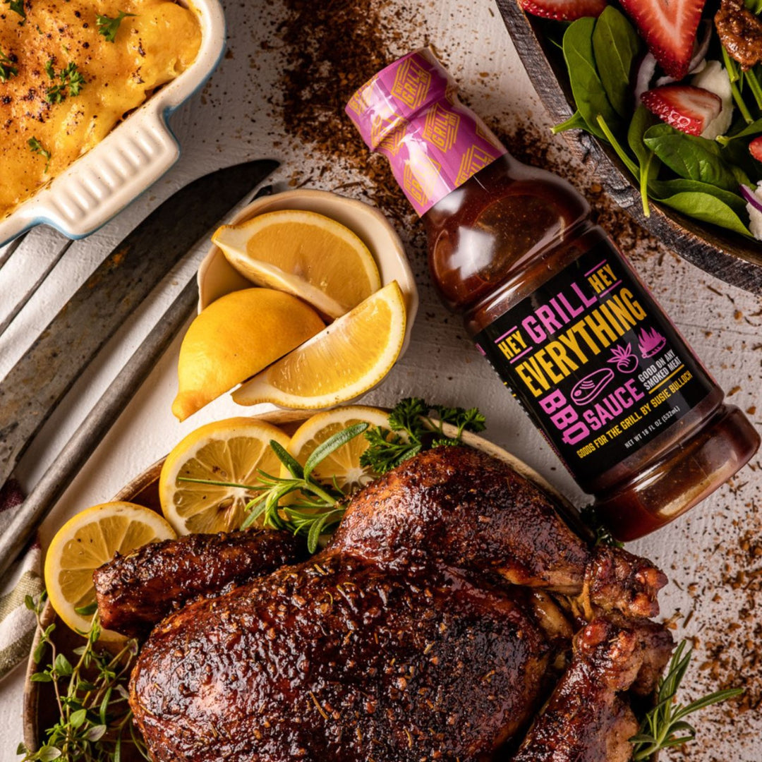 A bottle of Everything Sauce sits on a table next to a smoked whole chicken that has been plated with herbs and lemon. Other side dishes surround the area