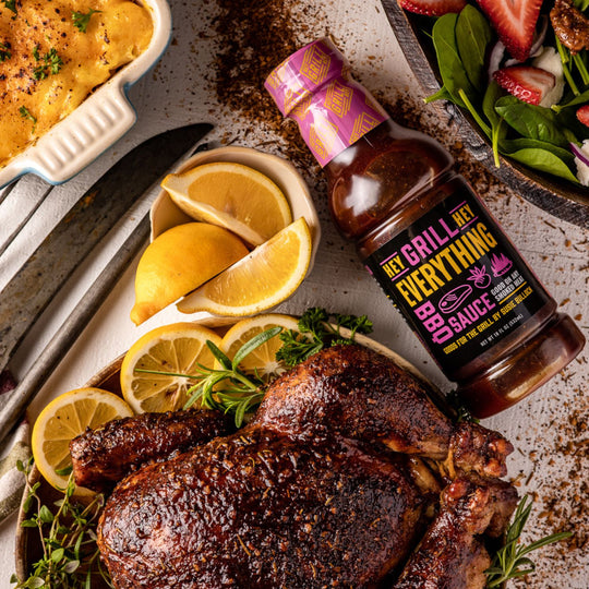 A smoked whole chicken sits plated with herbs and lemon and garnishing. A bottle of Everything BBQ Sauce sits on the table next to the chicken.  