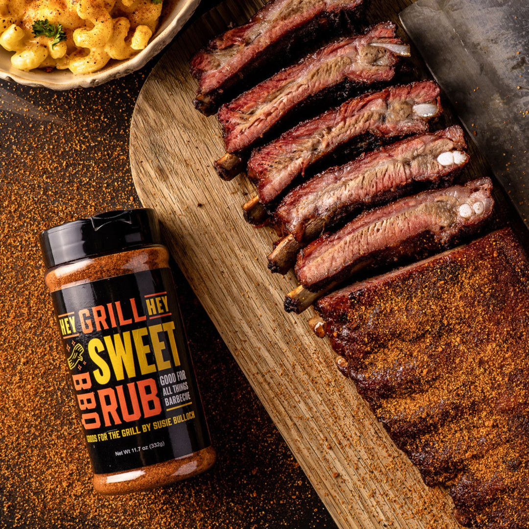 A bottle of sweet rub sits on a table next to a cut rack of smoked ribs, covered in Sweet Rub.