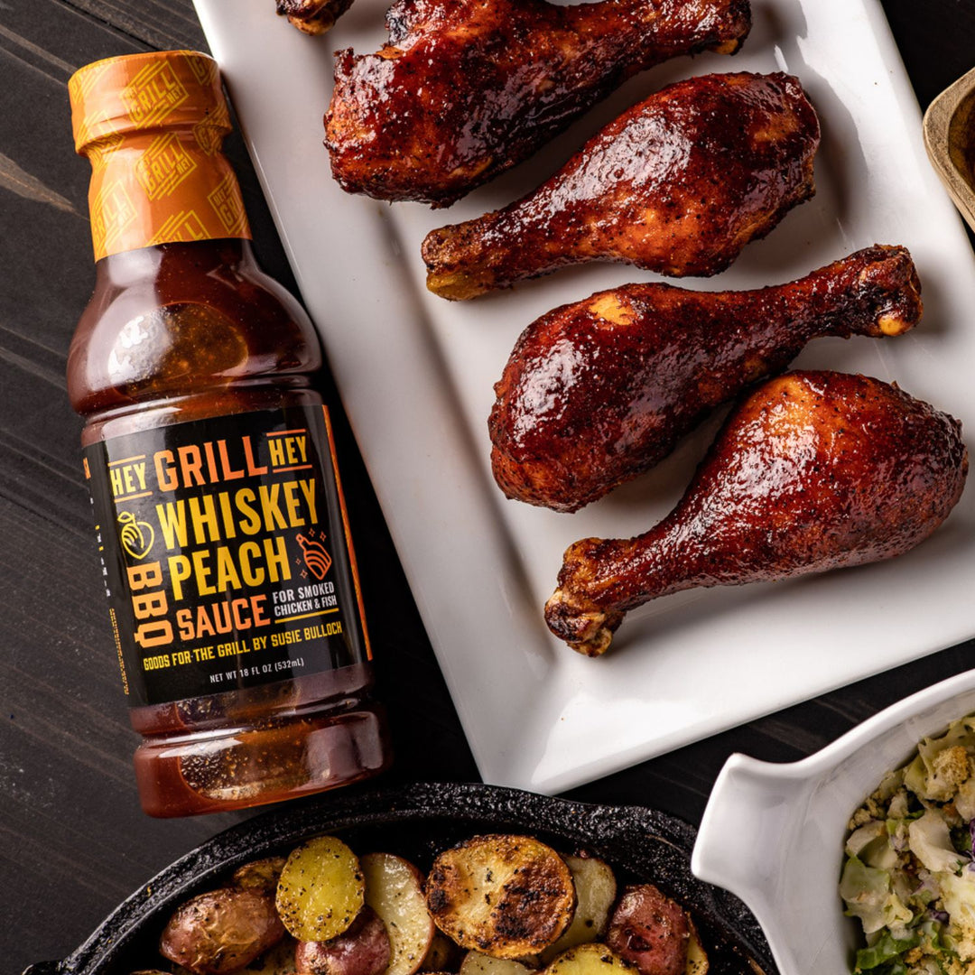 A bottle of Whiskey Peach Sauce sits on a table next to smoked chicken legs that have been covered in Whiskey Peach sauce. 